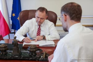 Na zdjęciu prezydent Karol Nawrocki w białej koszuli siedzący za biurkiem i podpisujący ustawę. Plecami do kadru patrzy na to Zbigniew Bogucki.