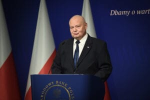 Prof. Adam Glapiński w garniturze w trakcie konferencji prasowej w siedzibie NBP. Za jego plecami widać biało-czerwone flagi na granatowym tle, zaś po prawej jest napis: "dbamy o wartość twojego pieniądza".