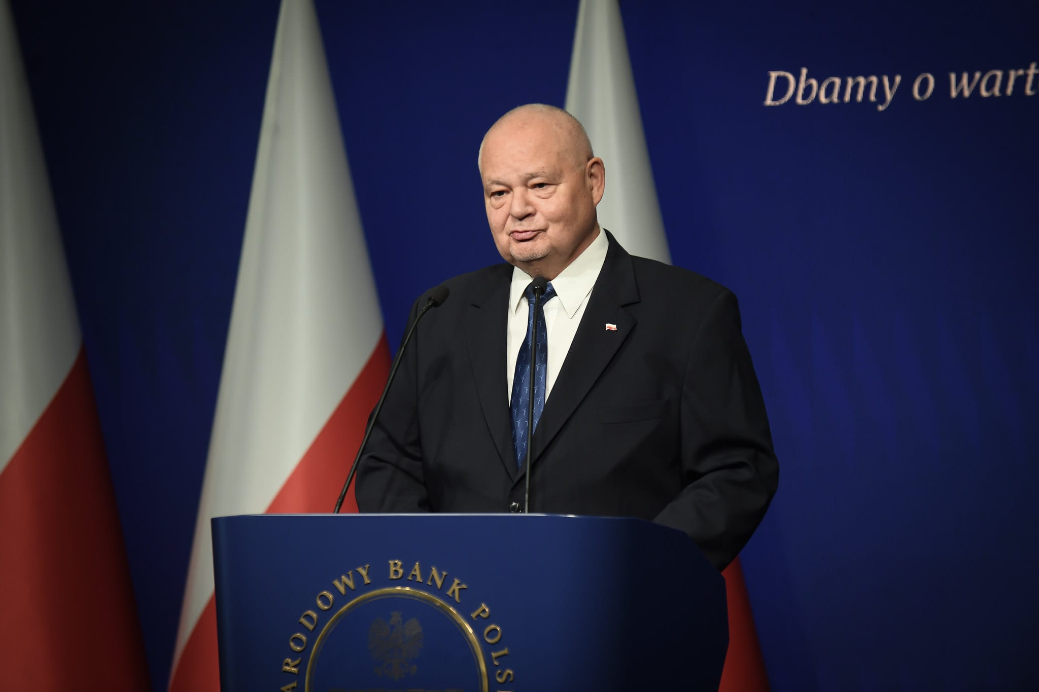 Prof. Adam Glapiński w garniturze w trakcie konferencji prasowej w siedzibie NBP. Za jego plecami widać biało-czerwone flagi na granatowym tle, zaś po prawej jest napis: "dbamy o wartość twojego pieniądza".