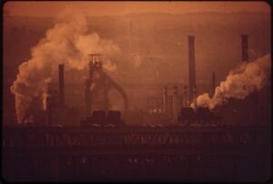 Pozioma fotografia przedstawiająca industrialny krajobraz o wschodzie słońca. Na pierwszym planie widoczne są czarne, kontrastowe sylwetki infrastruktury przemysłowej: wysokie kominy fabryczne, plątanina rur oraz słupy linii wysokiego napięcia. Tło stanowi zamglone niebo w odcieniach brudnego pomarańczu i szarości, przysłonięte gęstym dymem wydobywającym się z zakładu.