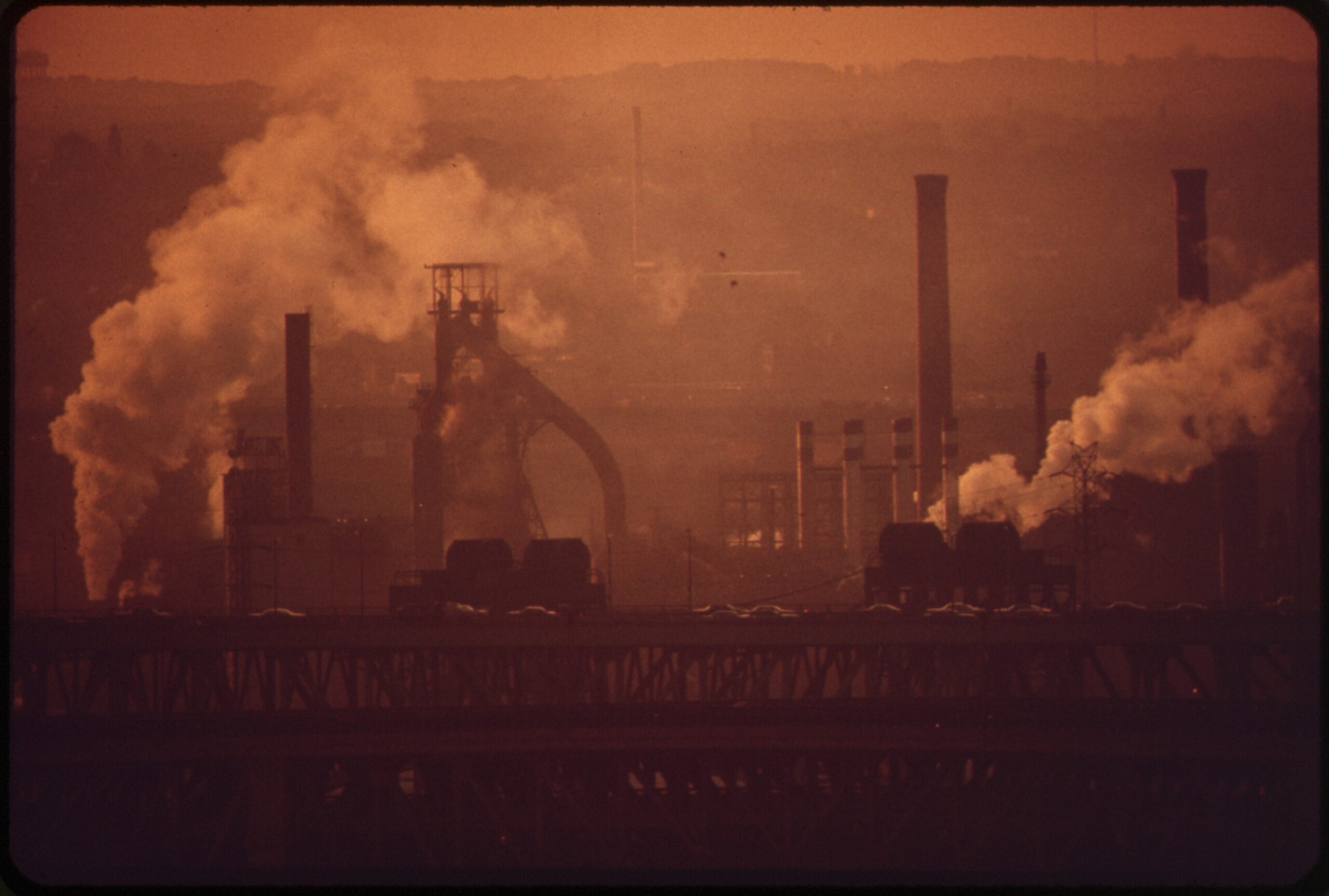 Pozioma fotografia przedstawiająca industrialny krajobraz o wschodzie słońca. Na pierwszym planie widoczne są czarne, kontrastowe sylwetki infrastruktury przemysłowej: wysokie kominy fabryczne, plątanina rur oraz słupy linii wysokiego napięcia. Tło stanowi zamglone niebo w odcieniach brudnego pomarańczu i szarości, przysłonięte gęstym dymem wydobywającym się z zakładu.