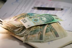 Banknoty o nominale 100 zł leżące na dokumentach. W tle widoczny długopis i zapisany formularz.