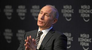 Władimir Putin w ciemnym garniturze i paskowanej krawacie siedzi na scenie ze złożonymi dłońmi, patrząc w górę na tle powtarzającego się logo World Economic Forum.