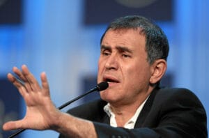 Nouriel Roubini w garniturze przemawiający do mikrofonu, z uniesioną dłonią i ekspresyjną mimiką.