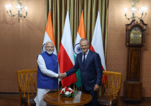 Premier Indii Narendra Modi i Donald Tusk podczas uścisku dłoni. Modi jest w tradycyjnym, indyjskim stroju, podczas gdy polski premier - w garniturze. W tle flagi obu państw.