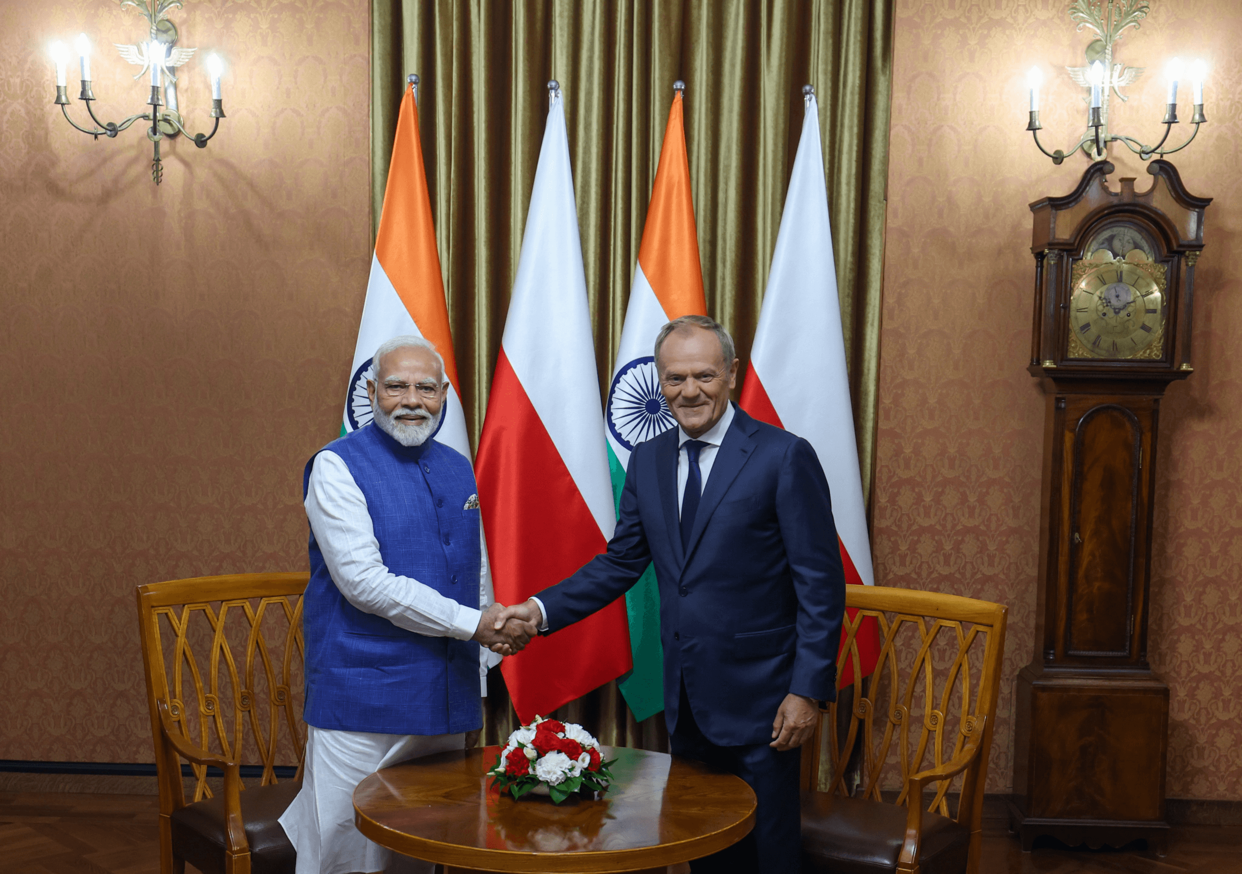 Premier Indii Narendra Modi i Donald Tusk podczas uścisku dłoni. Modi jest w tradycyjnym, indyjskim stroju, podczas gdy polski premier - w garniturze. W tle flagi obu państw.