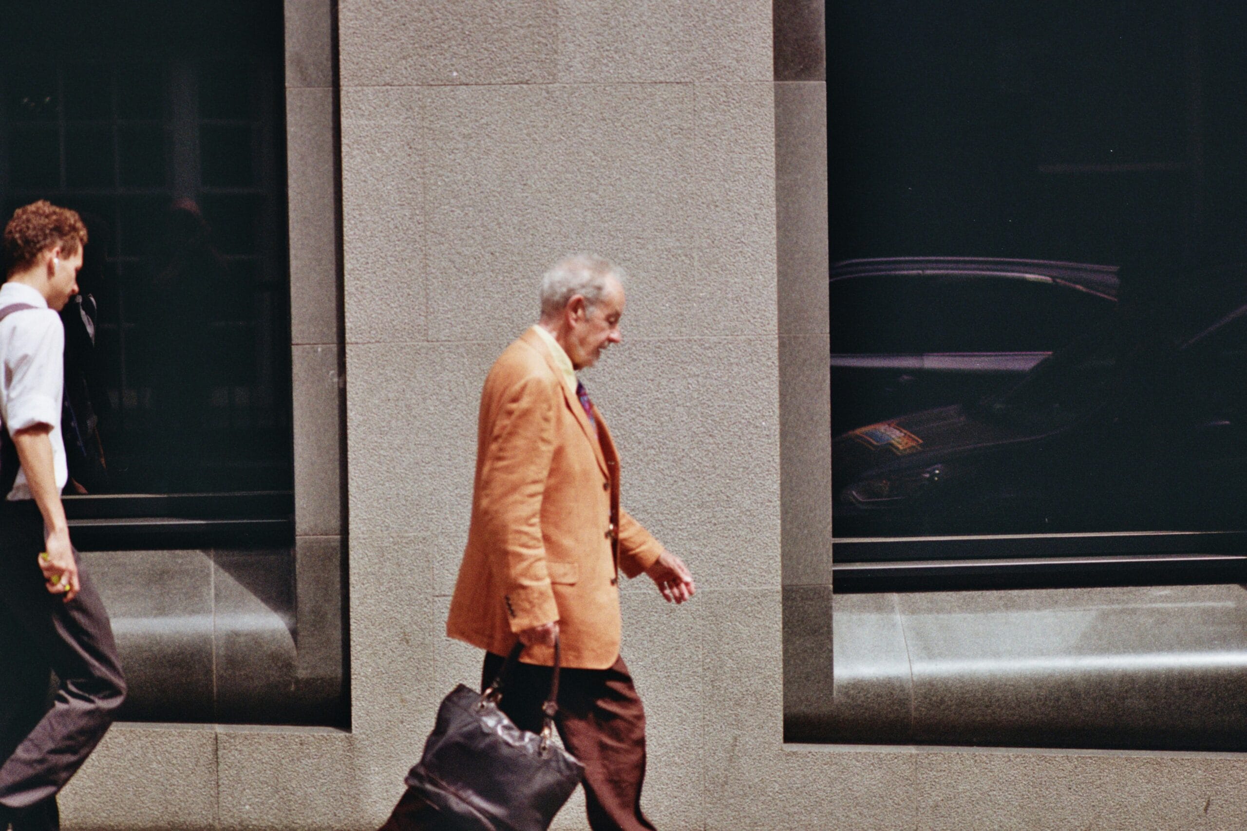 Starszy człowiek w pomarańczowej marynarce z teczką pod ręką idzie ulicą na tle biurowca. Za nim młodszy mężczyzna w koszuli z podwiniętymi rękawami.