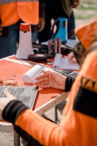 Robotnik w pomarańczowym kombinezonie siedzi przy stole. Obok ma laptopa i megafon