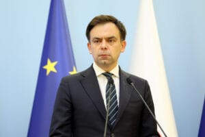 Andrzej Domański w garniturze stojący na mównicy w czasie konferencji prasowej. Za nim flagi Unii Europejskiej i Polski.