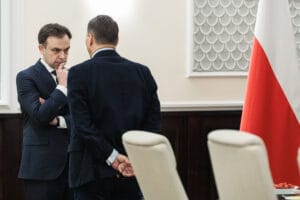 Andrzej Domański (stojący przodem do kamery) skupiony i pogrążony w rozmowie z Radosławem Sikorskim stojącym tyłem do obiektywu. Obaj panowie są w garniturach w sali, gdzie odbywają się posiedzenia rządu. Z prawej strony zdjęcia widać polską flagę.