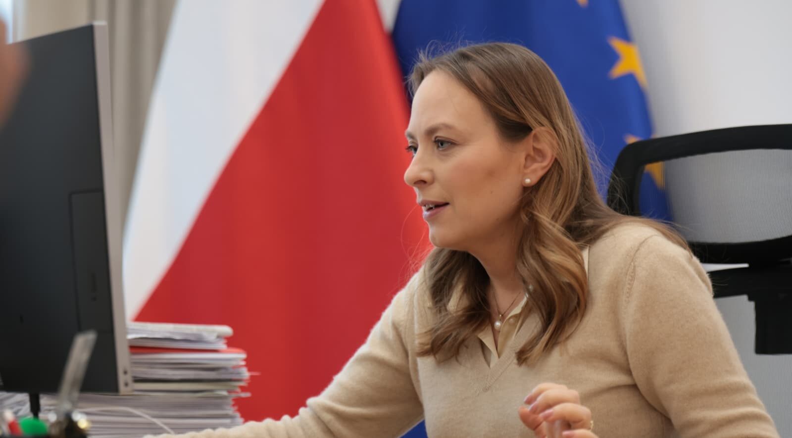 Katarzyna Pełczyńska-Nałęcz przy biurku z komputerem. Za nią znajdują się flagi polska i unijna