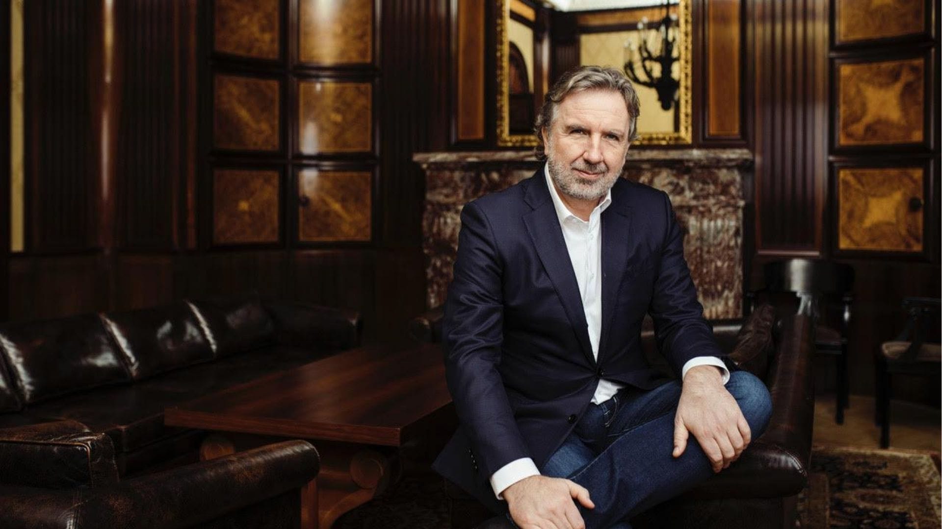 Siedzący Michał Sołowow w eleganckim garniturze na tle biblioteczki.