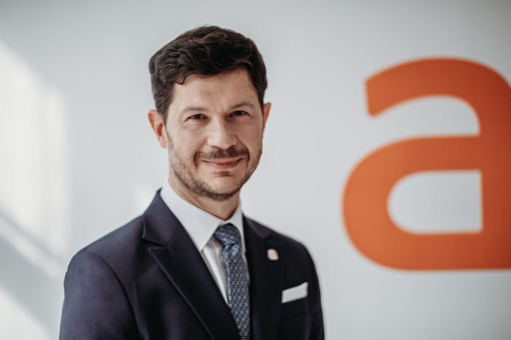 Marcin Kuśmierz w garniturze na tle białej ściany z pomarańczową literą "A", kojarzoną z logiem Allegro.