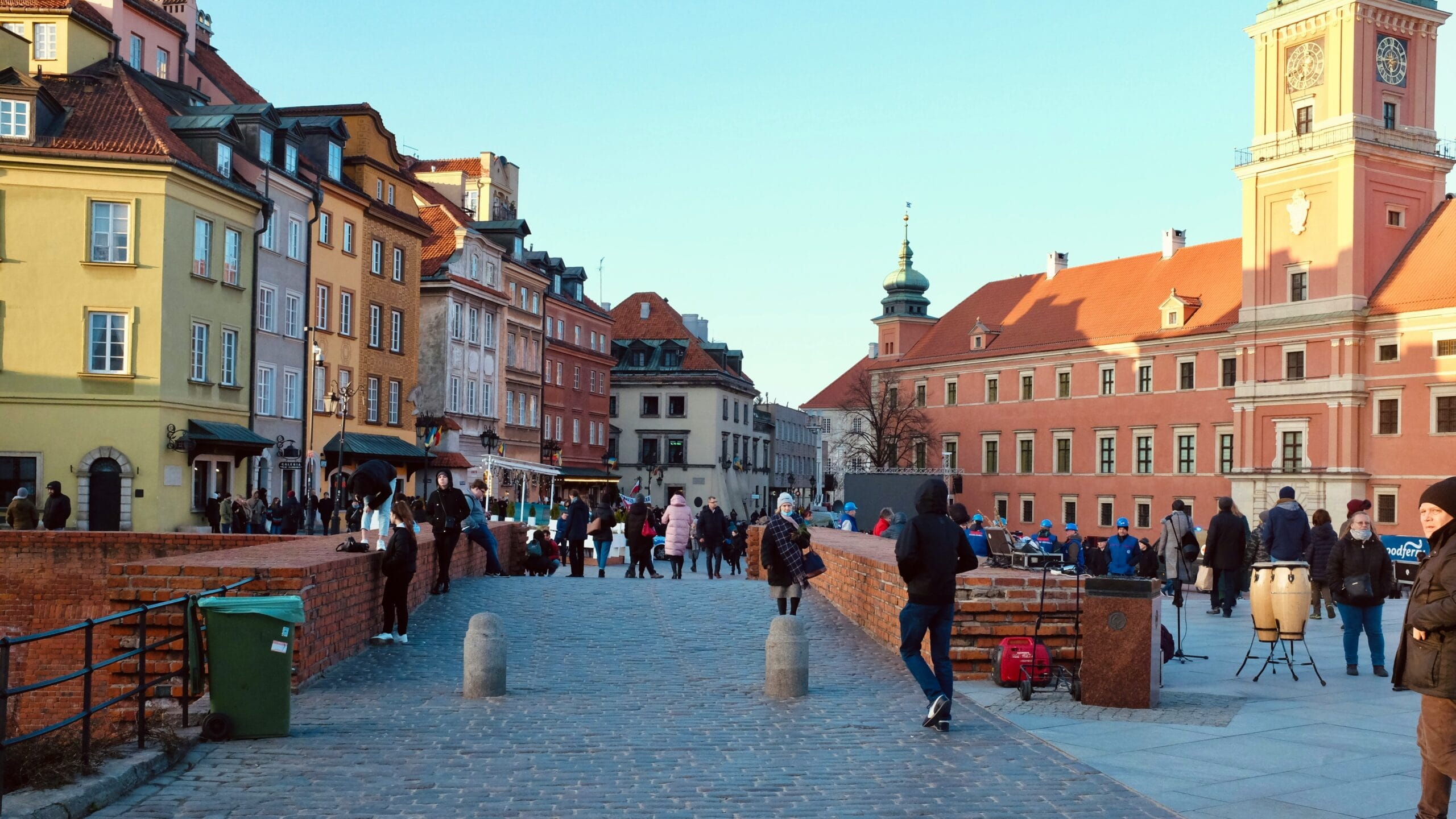 Ludzie spacerujący po Starym Mieście w Warszawie w aurze jesiennej; na dachu Zamku Królewskiego widać przebijające się promienie słoneczne.