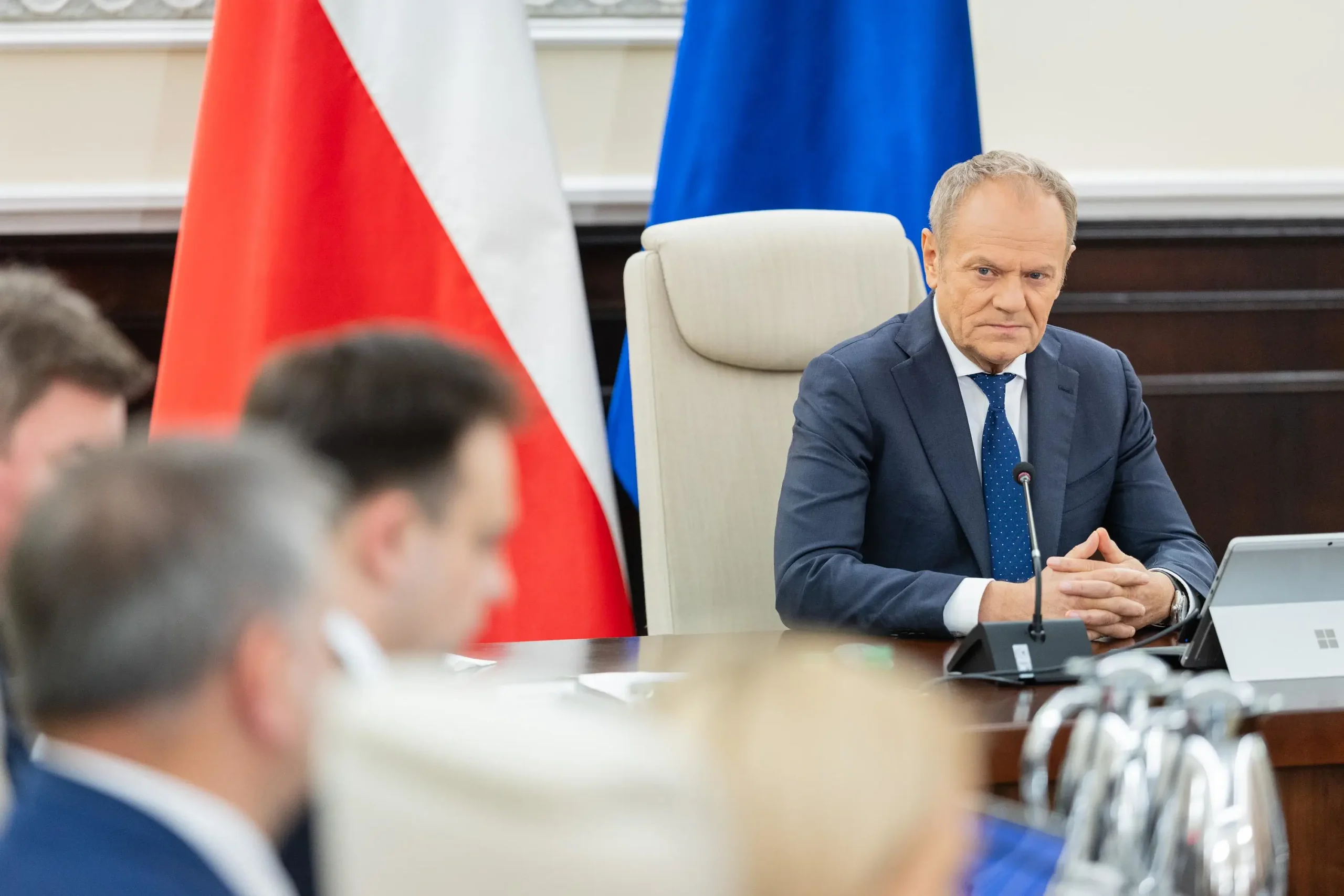 Donald Tusk podczas posiedzenia rządu. Premier wpatruje się z poważną miną w Andrzeja Domańskiego, który na zdjęciu jest rozmazany. Za plecami szefa rządu są polskie i unijne flagi.