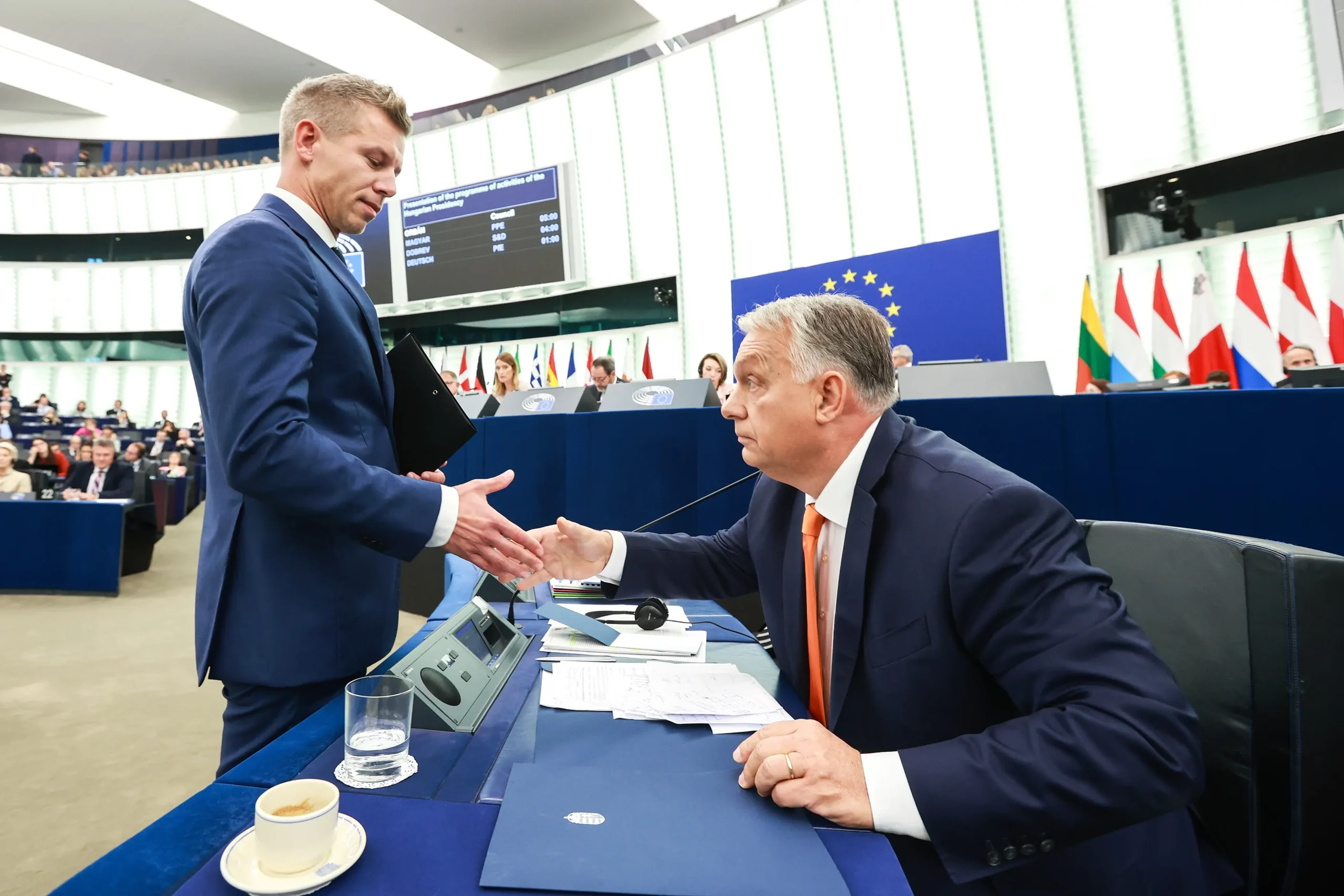 Stojący Péter Magyar podający rękę Viktorowi Orbánowi siedzącemu przy stole naprzeciw niego. Obydwaj w garniturach, spotkali się bowiem w Parlamencie Europejskim