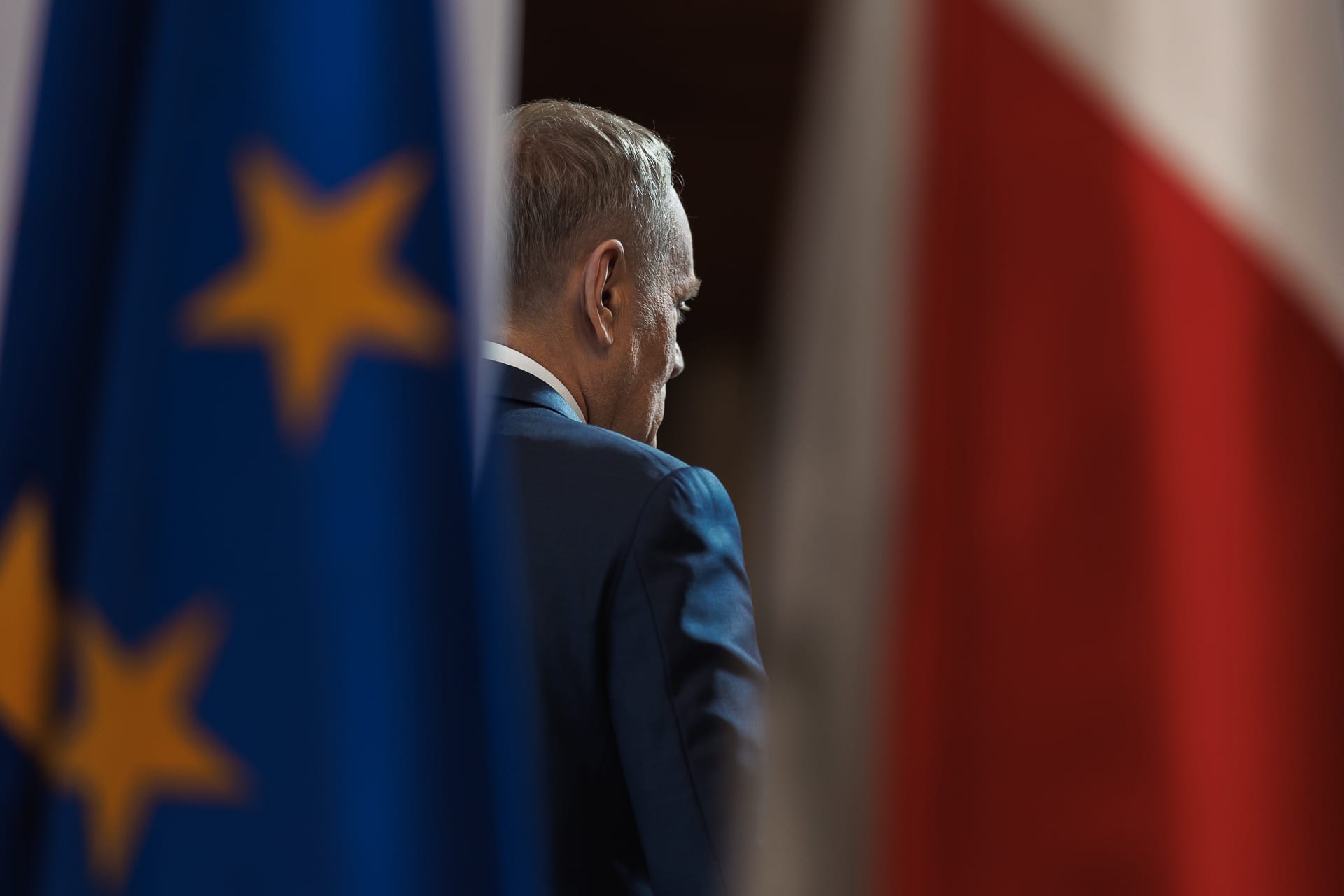 Odwrócony do kamery Donald Tusk cześciowo przysłonięty przez unijną flagę. Po jego prawej stronie znajduje się flaga Polski.