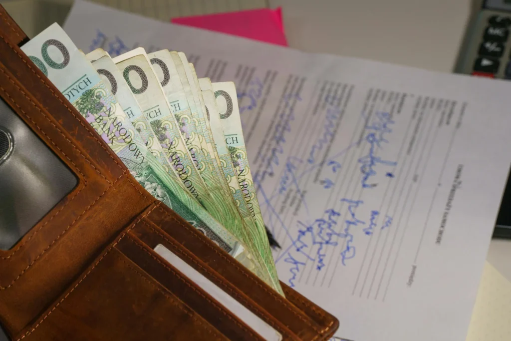 Brązowy portfel, z którego wystaje plik banknotów stuzłotowych na tle zapisanego niebieskim tuszem formularza.