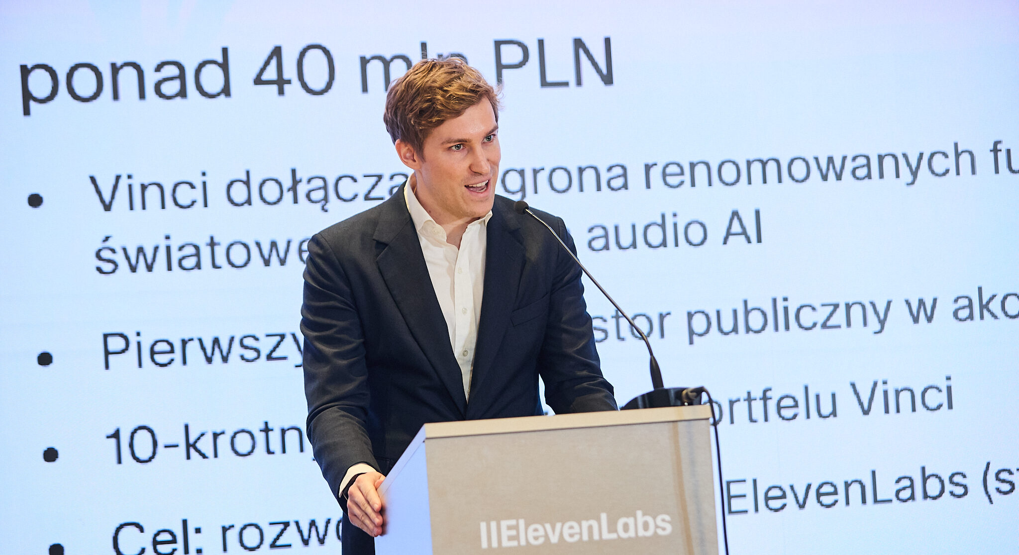 Mati Staniszewski na konferencji prasowej przy mównicy z napisem "ElevenLabs". Za nim slajd, na któym wybija się hasło "ponad 40 mln PLN"