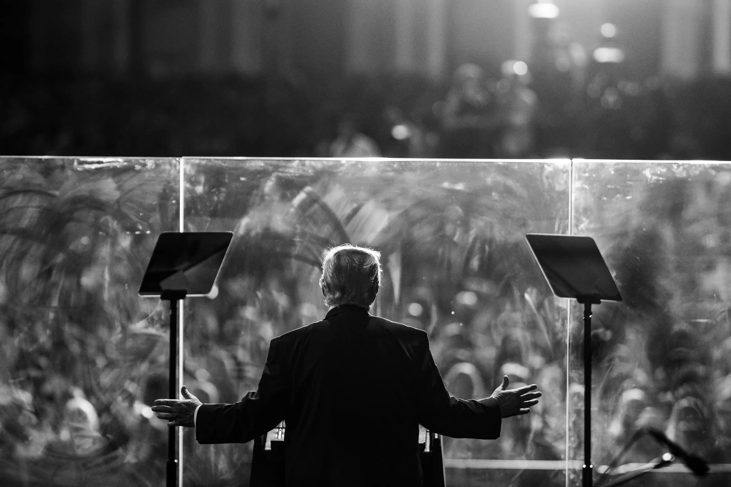 Stojący tyłem do obiektywu kamery Donald Trump w garniturze. Polityk skierowany jest do tłumu, do którego przemawia zza osłon pleksi.