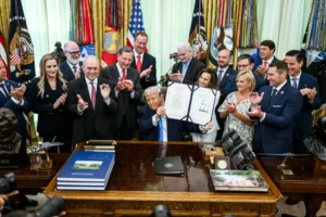 Zdjęcie przedstawia Donalda Trumpa trzymającego podpisaną ustawę w Gabinecie Owalnym